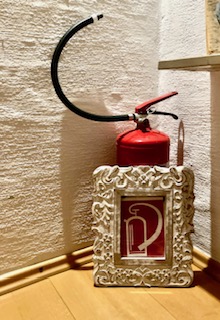 Roter Feuerlöscher mit schwarzem Schlauch steht in einer Ecke auf Holzboden, davor ein verziertes weißes Schild mit weißem Feuerlöscher-Symbol auf rotem Grund