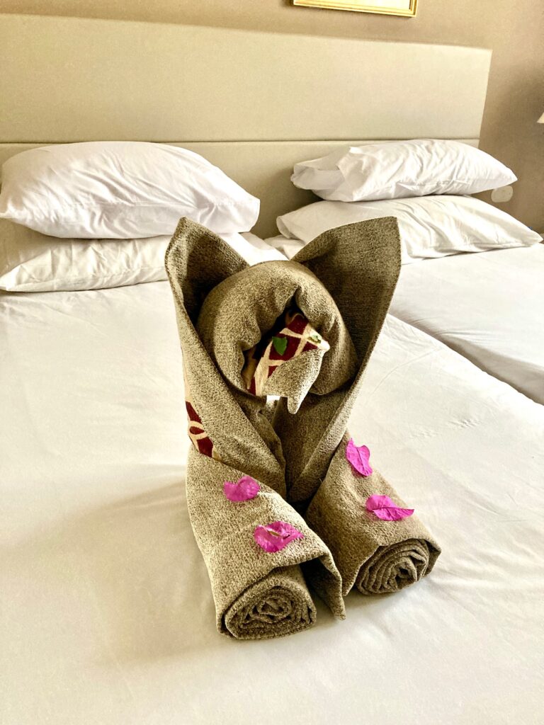 Zusammengerollter und gefalteter brauner Bettüberwurf auf einem Bett, geformt wie ein Tier mit Ohren, verziert mit rosa Blütenblättern