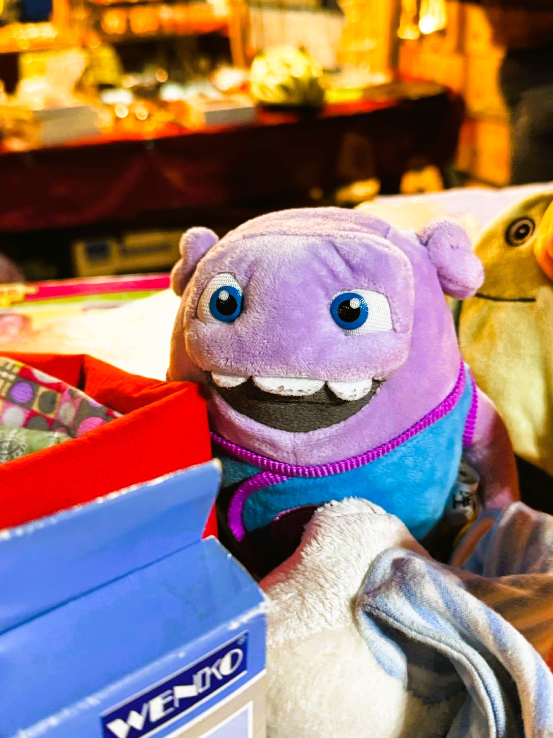 Lila Plüschfigur mit großen blauen Augen und breitem Lächeln, umgeben von anderen Stofftieren und Spielzeug, im Hintergrund ein nächtlich beleuchteter Flohmarktstand