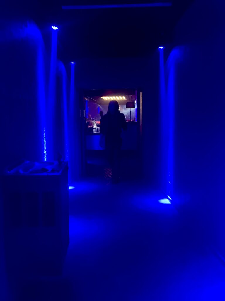 Dunkler Flur mit blauer Beleuchtung und einer Person im Hintergrund vor einer hell erleuchteten Bar