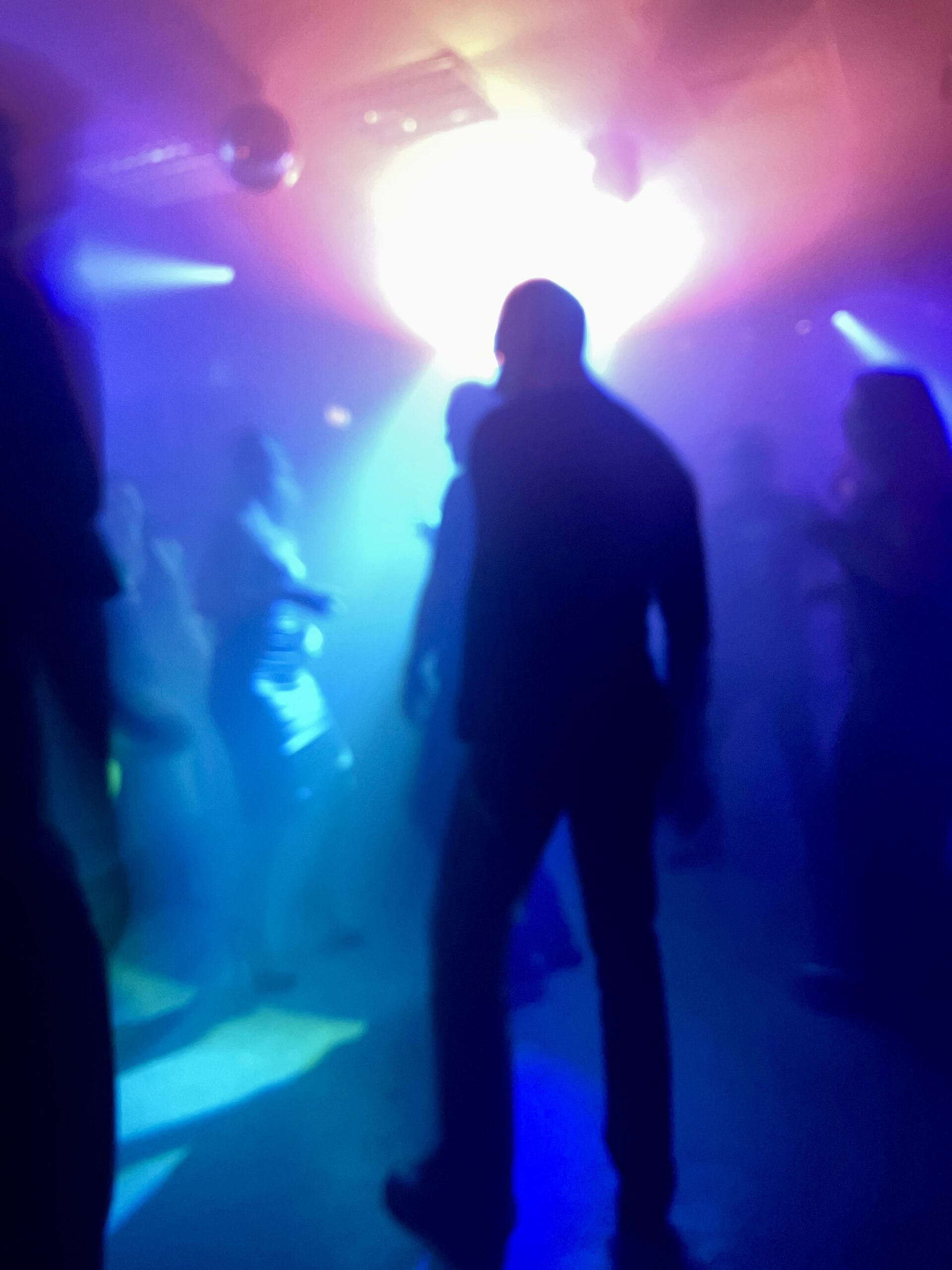 Silhouetten von tanzenden Personen im Schattenriss auf einer dunklen Tanzfläche mit blauem und gelbem Licht, im Vordergrund der Schattenriss einer tanzenden männlichen Person