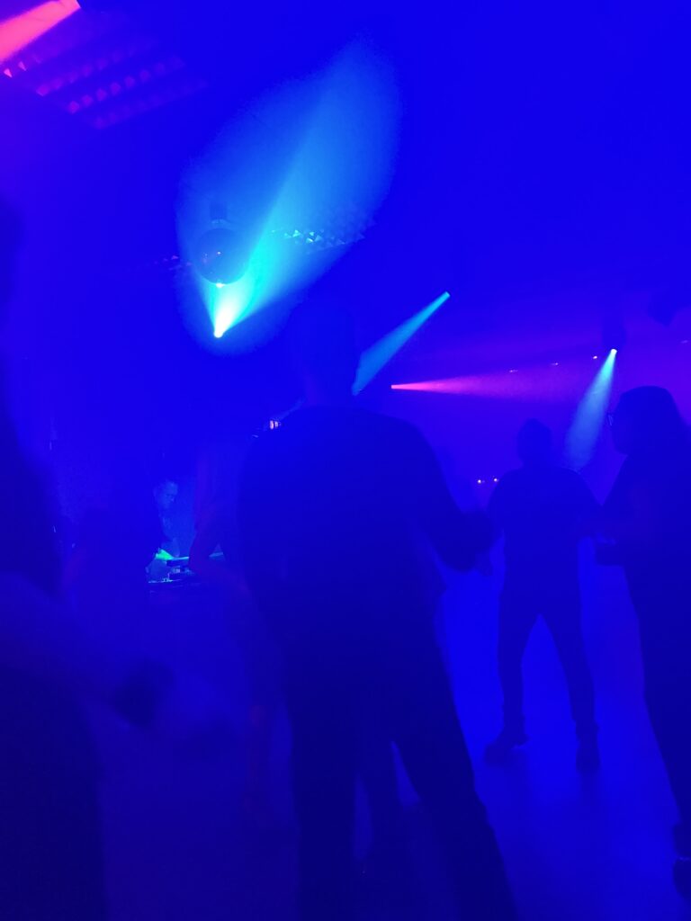 Tanzfläche mit tanzenden Menschen im Schattenriss in dunkler Umgebung mit blauem und violettem Bühnenlicht und Nebel