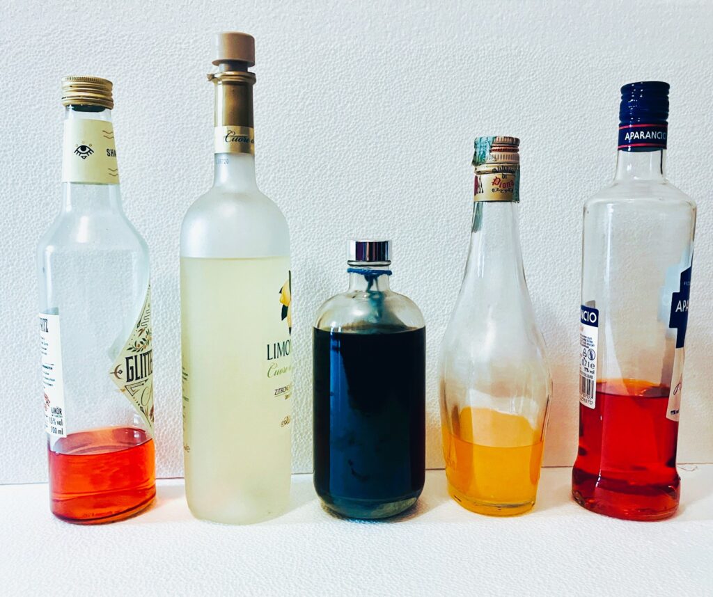 Fünf unterschiedlich volle Flaschen mit farbigen Flüssigkeiten in Rot, Hellgelb, Braun, Orange und Rot vor weißer Wand