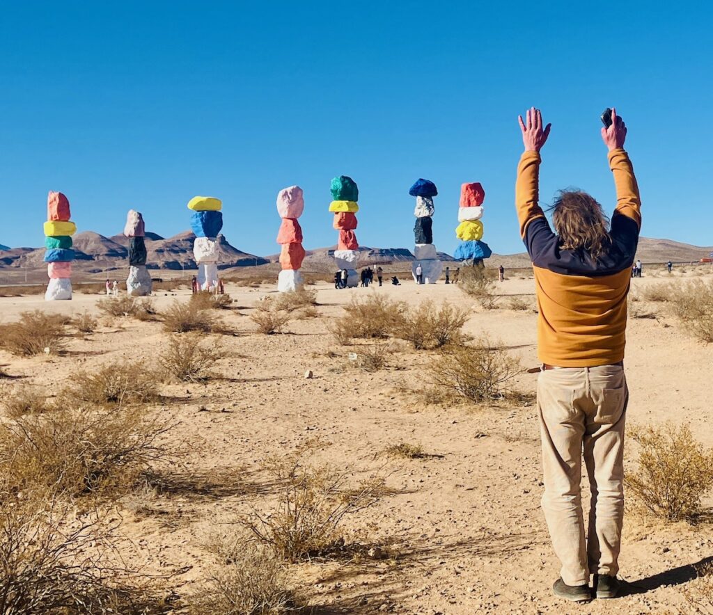 Person mit erhobenen Armen steht mit dem Rücken zum Betrachter vor mehreren hohen, bunt bemalten Steinsäulen in einer Wüstenlandschaft. Die Steine bilden die Kunstinstallation "Seven Magic Mountains" von Ugo Rondinone bei Las Vegas. Der Pullover der Person ist horizontal in die Farbflächen gelb und grau unterteilt