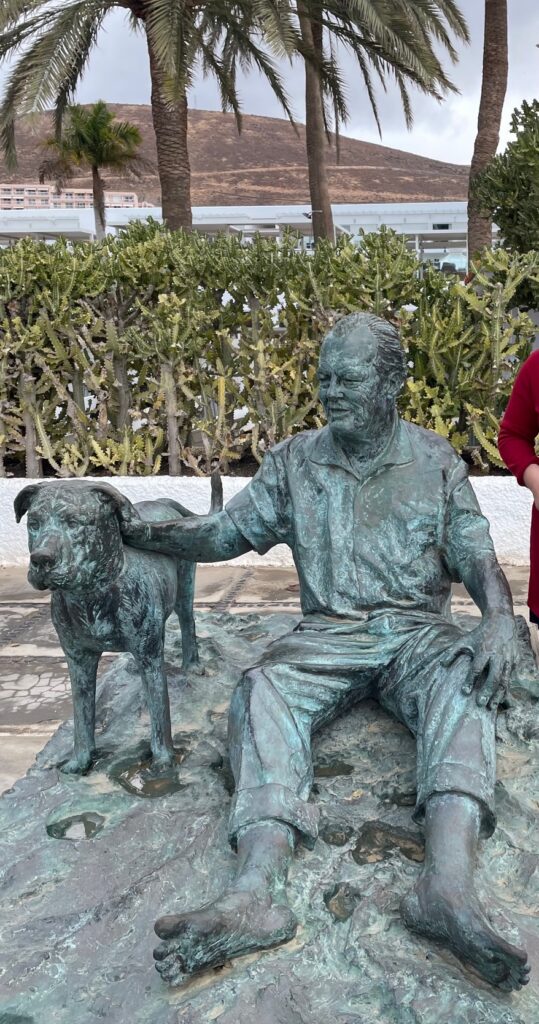 Lebensgroße Bronzestatue eines sitzenden Mannes mit einem Hund, umgeben von Kakteen und Palmen im Hintergrund. Die Statue zeigt Willy Brandt