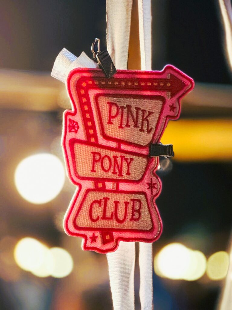 Stoffanhänger in Vintage-Pfeilform in rot und pink mit den Worten 'PINK PONY CLUB' befestigt an einem weißen Band, im Hintergrund verschwommene Lichter vor einem schwarzen Himmel