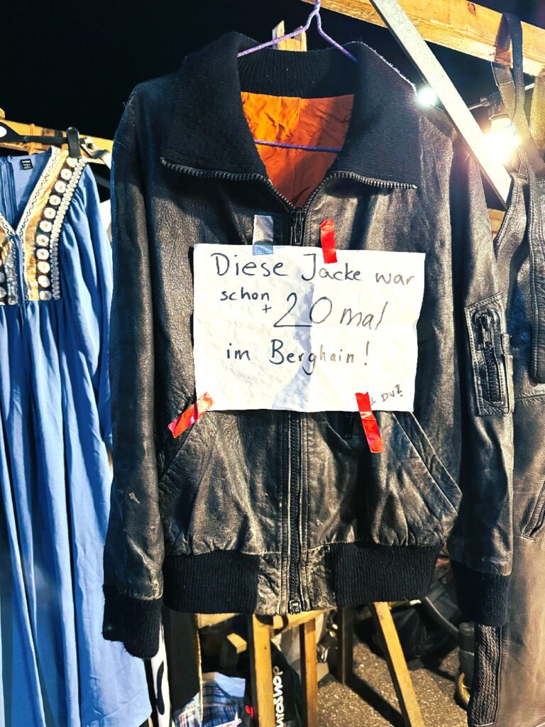 Schwarze Lederjacke mit Reißverschluss und orangefarbenem Innenfutter, daran befestigt ein handgeschriebener Zettel mit der Aufschrift 'Diese Jacke war schon 20 mal im Berghain!'