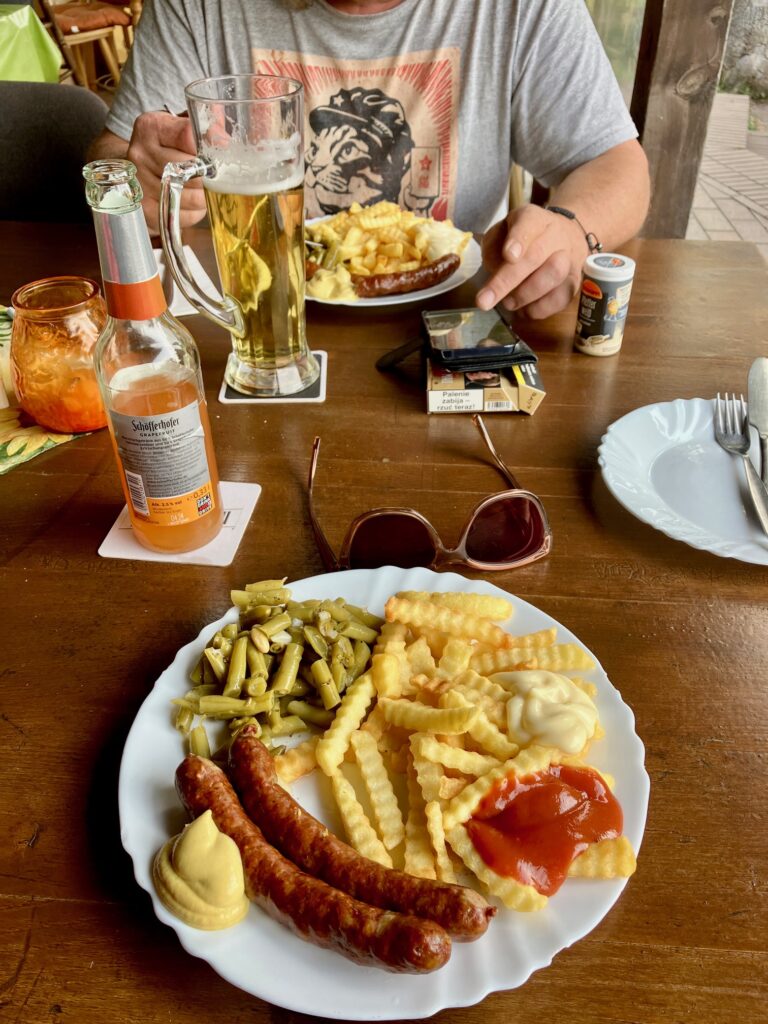 Teller mit Bratwürsten, Senf, grünen Bohnen, Pommes mit Mayonnaise und Ketchup auf Holztisch, im Hintergrund Person mit Bierglas und Teller mit Pommes