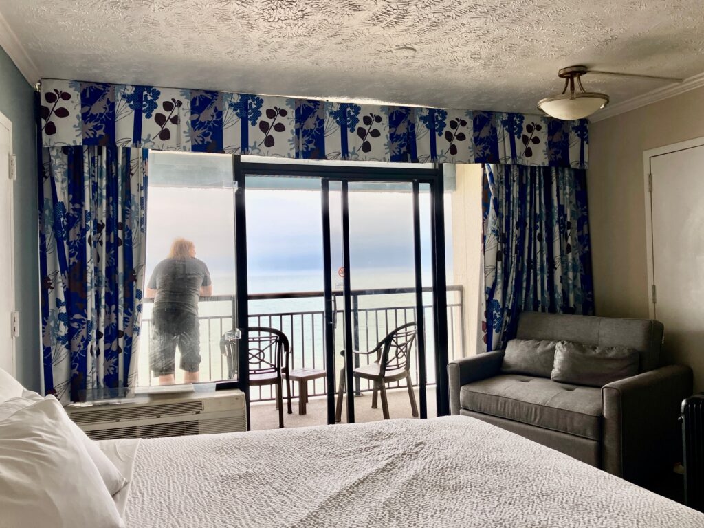 Hotelzimmer mit Bett, Sofa und Balkon, auf dem eine Person steht und aufs Meer blickt