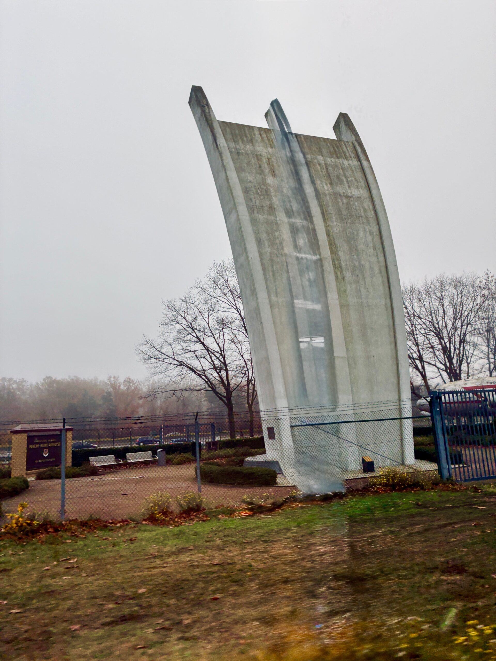 Eine hohe, gebogene Betonfläche mit drei vorstehenden Stäben steht auf einem umzäunten Gelände unter weißem Himmel