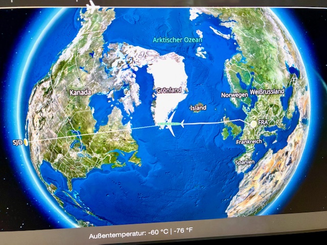 Bildschirm mit Weltkugel im Flugzeug, darauf zu sehen ist Nordatlantik, Grönland, Kanada, Island, Norwegen, Russland. Eine Flugroute ist eingezeichnet, ein symbolisches Flugzeug ist an der Südspitze Grönlands zu sehen. Dazu Text 'Außentemperatur: -60 °C | -76 °F'