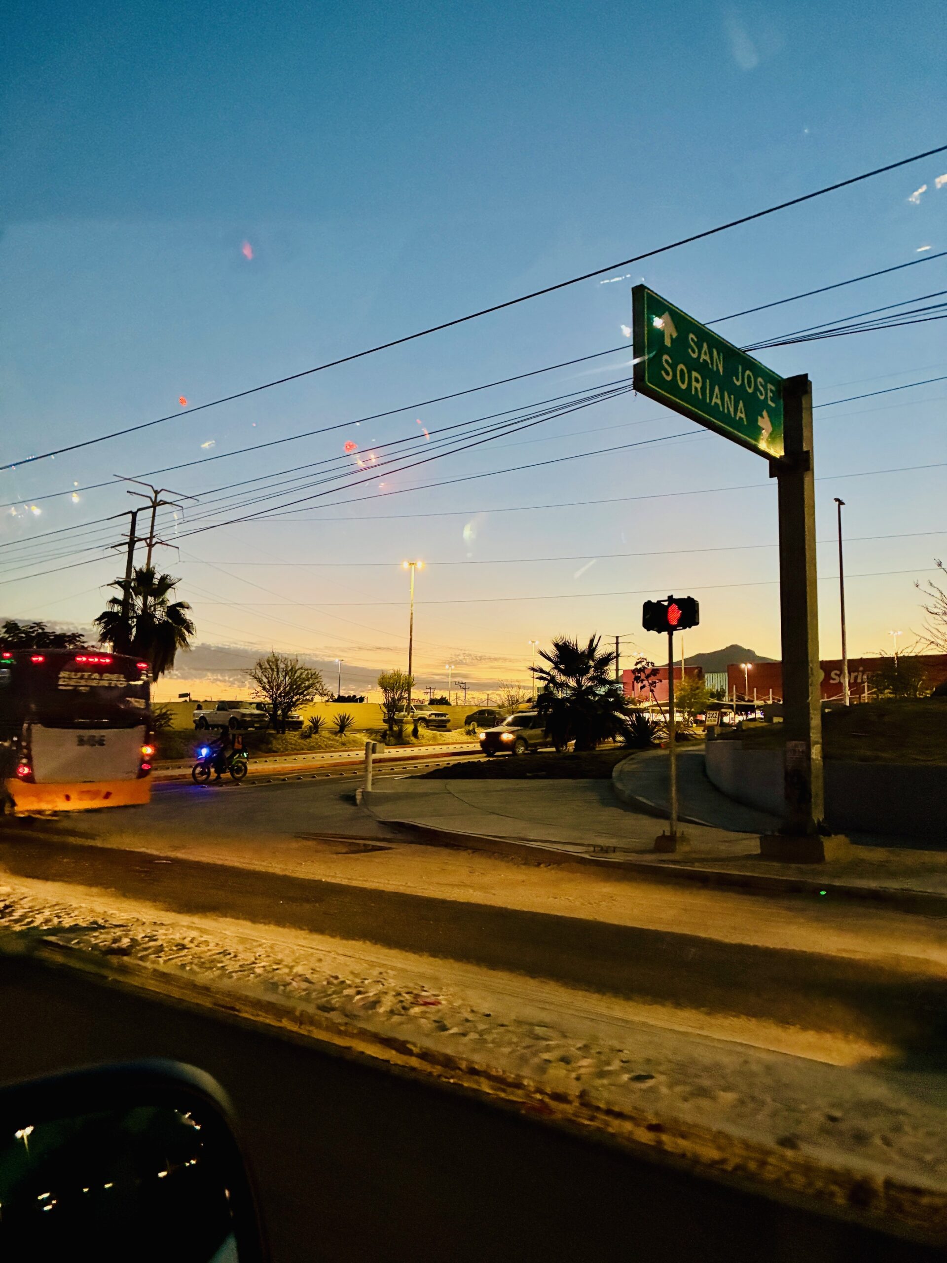 Straßenszene bei Abenddämmerung mit grünem Verkehrsschild zu San Jose und Soriana, Bus und Fahrradfahrer sichtbar