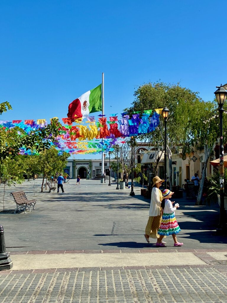 Eine Frau und ein Kind gehen über einen Platz mit bunten Papiergirlanden und einer mexikanischen Flagge unter tiefblauem Himmel. Das Kleid des Kindes hat die selben Farben wie die Papiergirlanden
