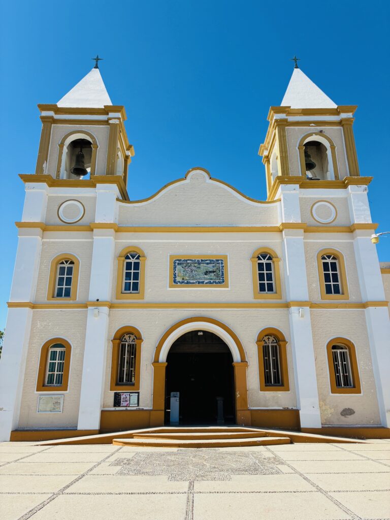 Fassade einer weißen Kirche mit zwei eckigen Glockentürmen, gelben Umrandungen und blauem Himmel im Hintergrund. Es handelt sich um die Misíon San José del Cabo