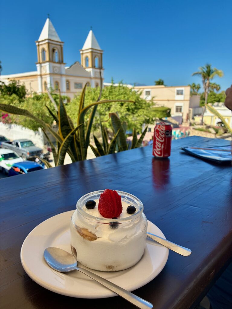 Glas mit Dessert, garniert mit einer Erdbeere und zwei Heidelbeeren, auf weißem Teller mit Löffeln auf Holztisch, im Hintergrund Kirche und Coca-Cola-Dose unter tiefblauem Himmel