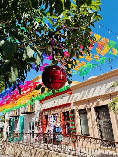 Rote Weihnachtskugel hängt an einem Baumzweig vor einem Haus mit bunten Papiergirlanden und blauem Himmel