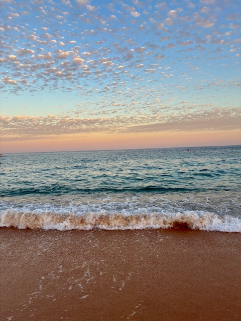 Strand mit sanften Wellen, darüber Himmel mit verstreuten kleinen Wolken bei Sonnenuntergang. Das Wasser mit weißer Gischt glitzert metallisch türkis, der Himmel ist hellblau und rosa