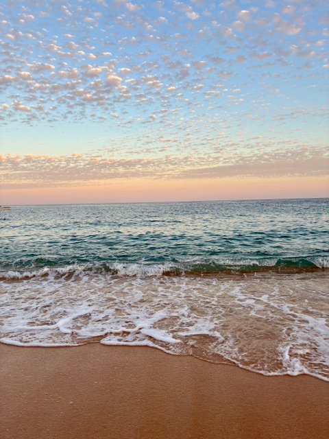 Strand mit sanften Wellen und leichter Brandung, darüber Himmel mit verstreuten kleinen Wolken bei Sonnenuntergang. Das Wasser mit weißer Gischt glitzert metallisch türkis, der Himmel ist hellblau und rosa