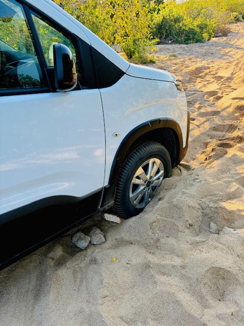 Vorderer Teil eines weißen Vans mit feststeckendem Vorderrad im Sand, umgeben von kleinen Steinen und grüner Vegetation