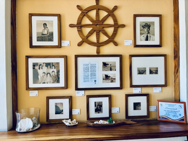Wand mit neun gerahmten Schwarz-weiß-Fotos und einem hölzernen Schiffssteuerrad in der Mitte, darunter ein Holztisch mit Glasgefäß und Muscheln