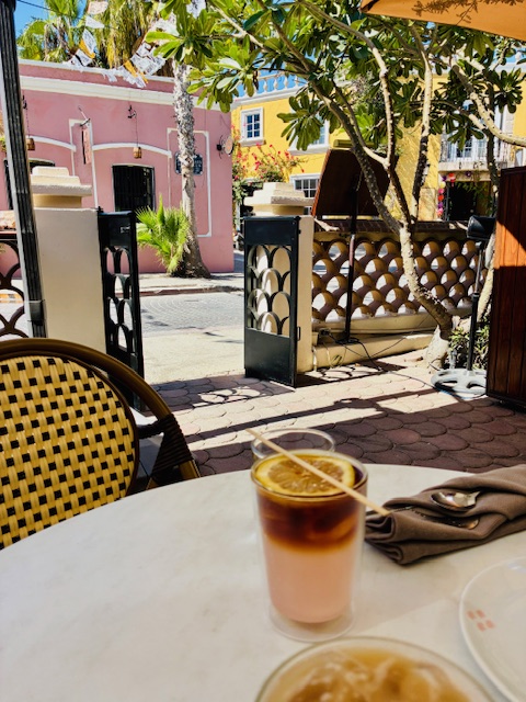 Trinkglas mit zweischichtigem Getränk, auf dem eine Zitronenscheibe liegt, auf weißem Tischtuch, daneben Besteck in Serviette, im Hintergrund bunte Häuser und Schatten von Bäumen.