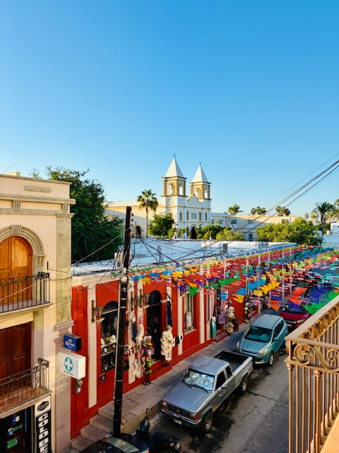 Blick von einem Balkon auf eine Straße, die mit bunten Papiergirlanden überspannt ist. Rote Häuser, zwei geparkte Autos und eine weiße Kirche mit zwei Türmen im Hintergrund unter blauem Himmel