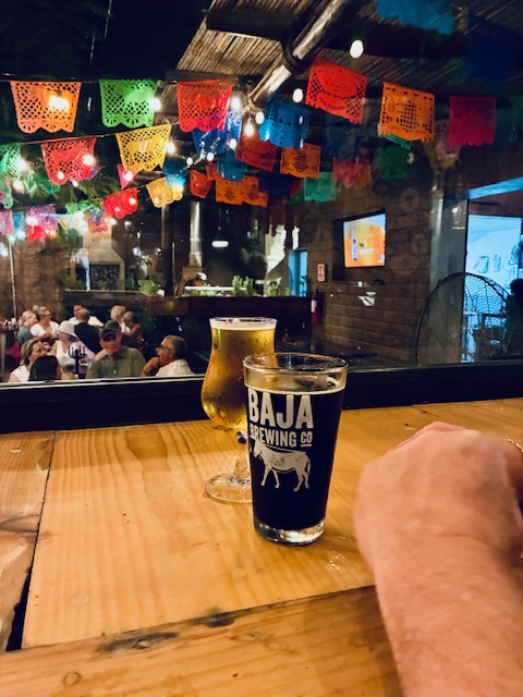 Zwei Biergläser auf einer Holztheke, eines mit dunklem Bier und Aufdruck 'BAJA BREWING CO', bunte Papiergirlanden im Hintergrund