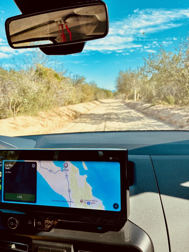 Blick aus einem Auto auf eine staubige, von Büschen gesäumte Landstraße unter blauem Himmel, im Armaturenbrett ein Navigationsgerät mit Kartenanzeige, auf dem der Nördliche Wendekreis am aktuellen Standort eingezeichnet ist