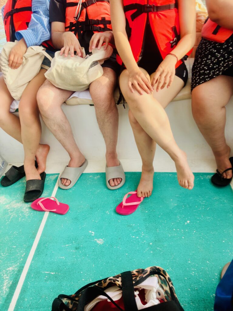 Vier Personen sitzen nebeneinander auf einer Bank, tragen orangefarbene Schwimmwesten und haben unterschiedliche Schuhe oder Flipflops an oder aus, darunter pinke Flipflops und graue Hausschuhe. Man sieht nur die Beine und Oberkörper der Personen und den türkisfarbenen Boden