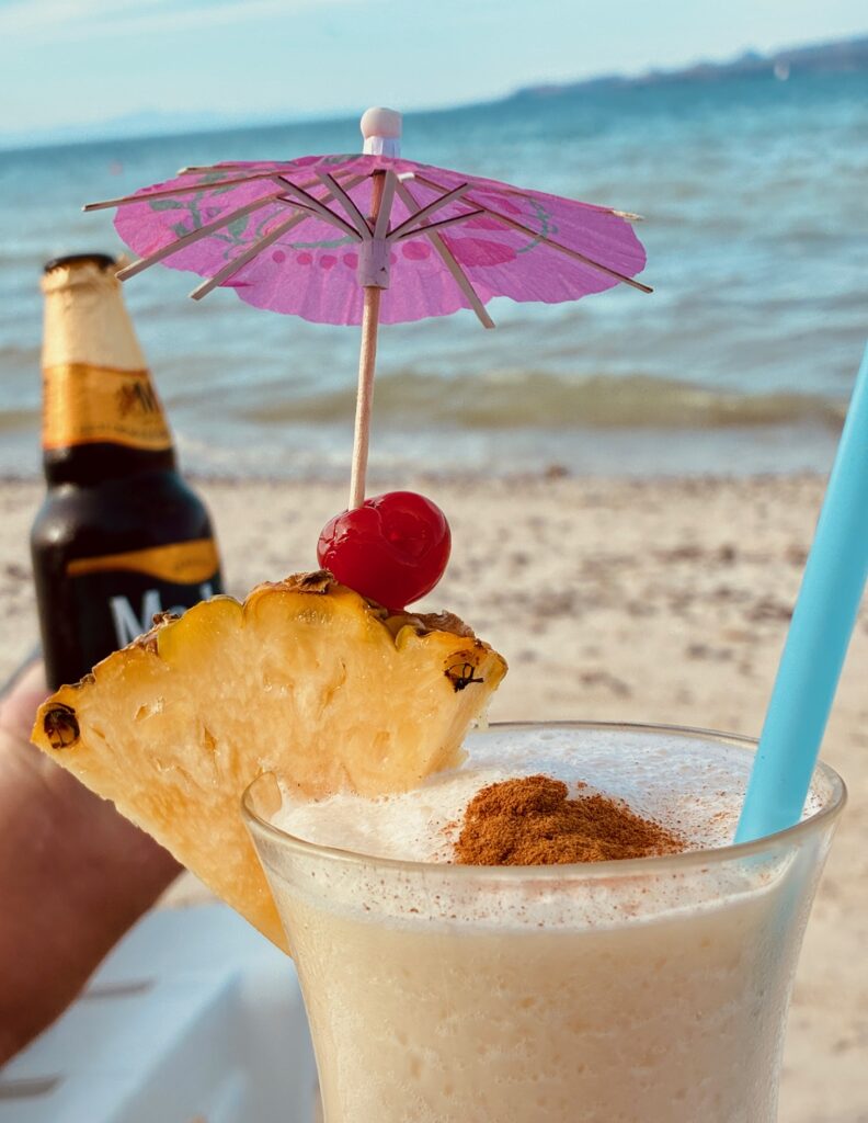 Cocktail mit Ananasscheibe, Cocktailkirsche und rosa Papierschirm am Strand, im Hintergrund eine Bierflasche und das Meer