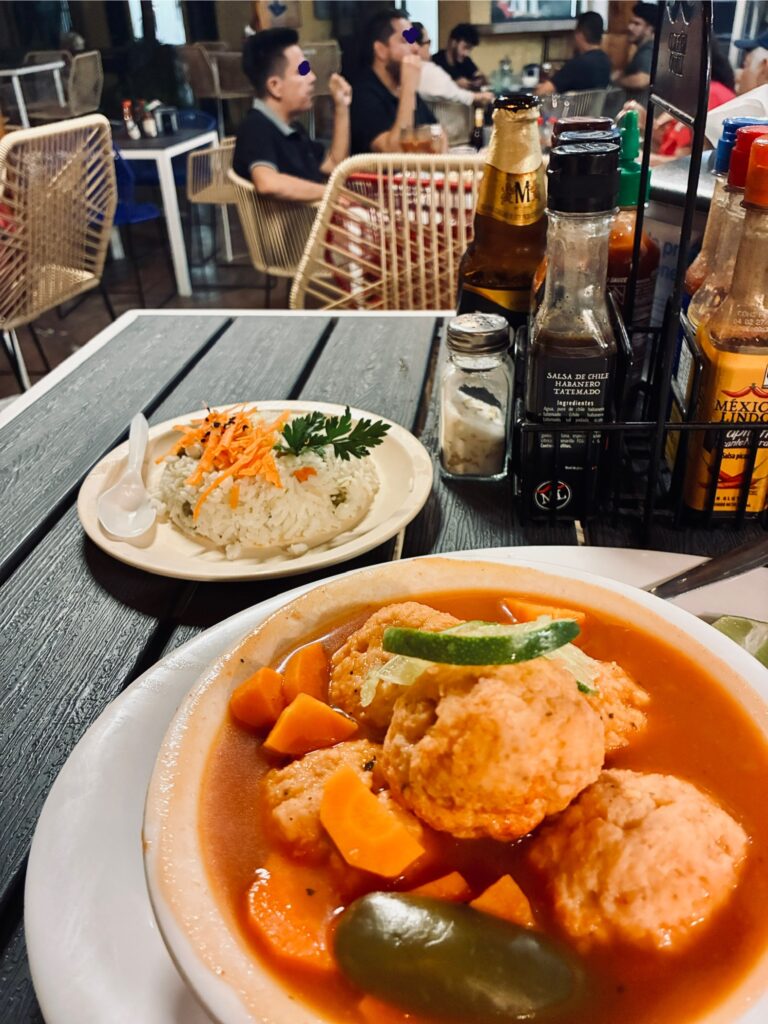 Teller mit Fischbällchen in roter Sauce, Karottenstücken und Limettenscheibe auf Holztisch, im Hintergrund Gewürzflaschen und Gäste in Restaurant