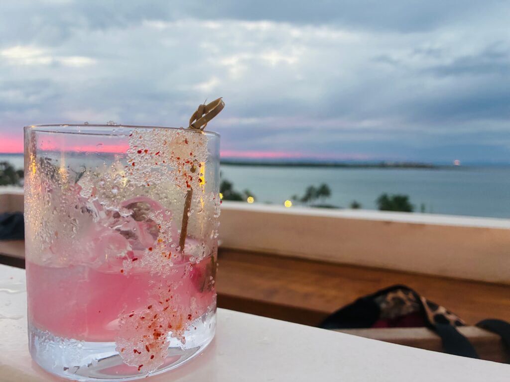 Glas mit rosa Getränk, Eiswürfeln und Salzdekoration auf einer weißen Brüstung, im Hintergrund Meer und bewölkter Himmel bei rosa Sonnenuntergang