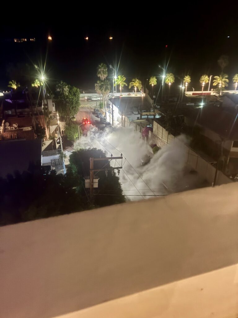 Nachtaufnahme einer Straße mit mehreren hellen Straßenlaternen, Palmen und Nebel oder Rauch, der sich über die Straße ausbreitet