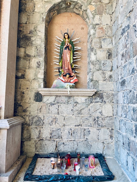 Statue der Jungfrau Maria mit grünem Umhang und rosafarbenem Kleid in einer Nische aus grauem Stein, darunter Kerzen auf dem Boden