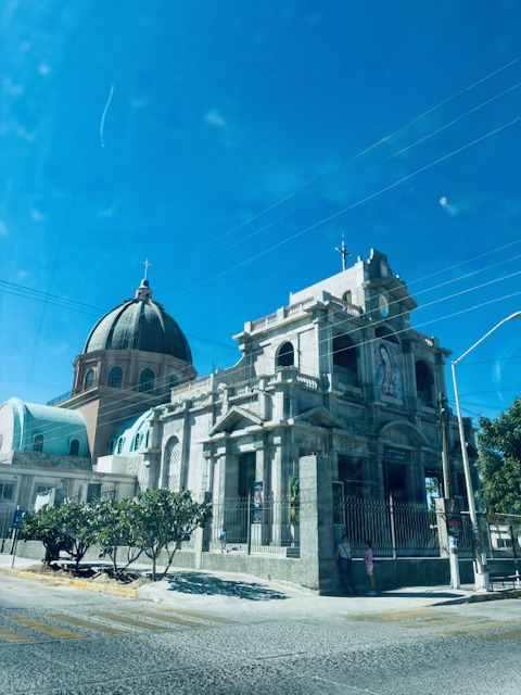 Großer Dom mit Kuppel an einer Straßenecke unter blauem Himmel, Bäume und zwei Personen am Zaun sichtbar