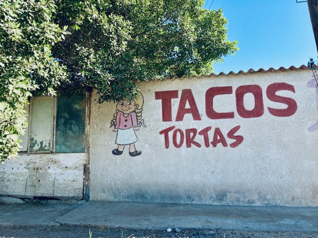 Wand mit roter Aufschrift 'TACOS TORTAS' und einer gemalten Figur eines Mädchens mit Zöpfen, rosa Jacke und weißem Rock neben einer alten Holztür unter blauem Himmel