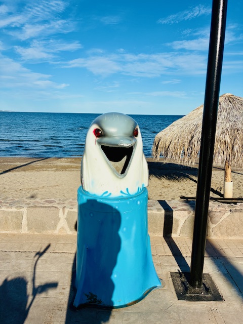 Blau-weißer Müllbehälter in Form eines Delfinkopfes mit offenem Maul, die Augen wurden rot besprüht, am Strand mit Meer und Strohhütte im Hintergrund unter blauem Himmel