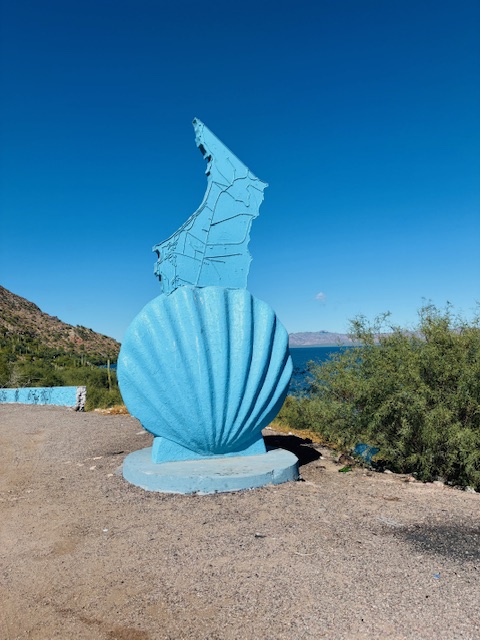 Blau gestrichene Skulptur mit einer großen Muschel und einer Landkarte, im Hintergrund ee und bewachsene Hügel.