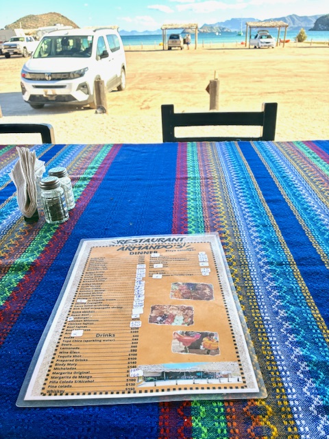 Bunt gestreifte Tischdecke mit Speisekarte eines Restaurants, im Hintergrund ein weißer Van und Strand mit Hütten