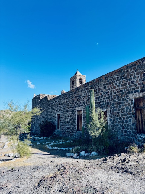 Historisches Gebäude aus dunklen Steinen mit kleinen Fenstern und einem Turm unter blauem Himmel in einer trockenen Landschaft mit Kakteen bei blauem Himmel