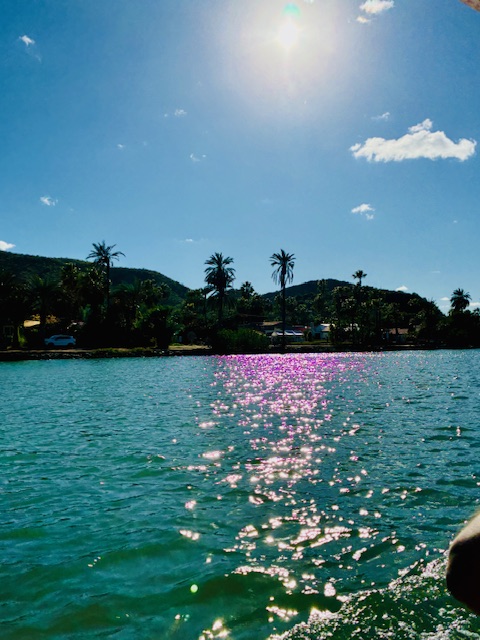 Sonnenlicht spiegelt auf türkisfarbenem Wasser, im Hintergrund Hügel mit Palmen und vereinzelten Häusern