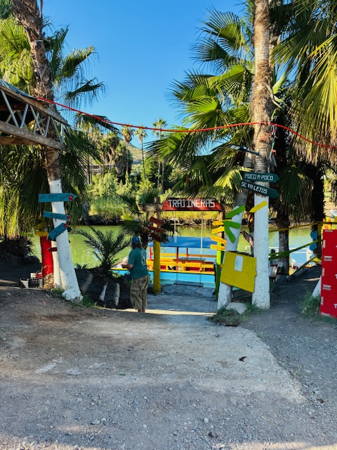 Eingang zu einem Bootsanleger mit bunten Holztoren, Palmen und einem Mann in blauer Mütze und grüner Hose, der auf das Wasser blickt