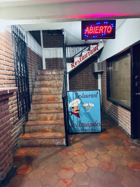 Treppenaufgang mit braunen Fliesen, links Backsteinwand mit vergittetem Fenster, an der Decke eine rote LED-Anzeige 'ABIERTO' und neben der Treppe ein altmodisches, hellblaues Schild mit Aufschrift 'Restaurant Los Equipales Bienvenido' und den Zeichnungen eines Kochs und zweier Cocktails