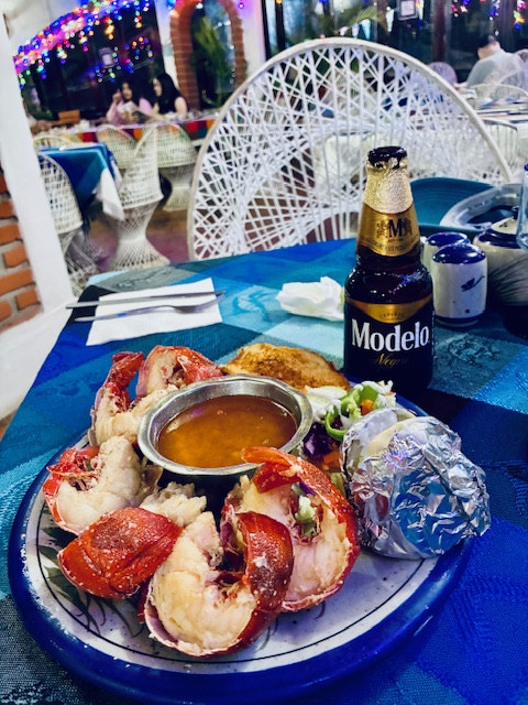 Teller mit großen Garnelen, einer kleinen Schale Soße, Folienkartoffel und Beilage, daneben eine Flasche Modelo-Bier auf blauem Tisch, dahinter Lichterketten und andere Tische des Restaurants