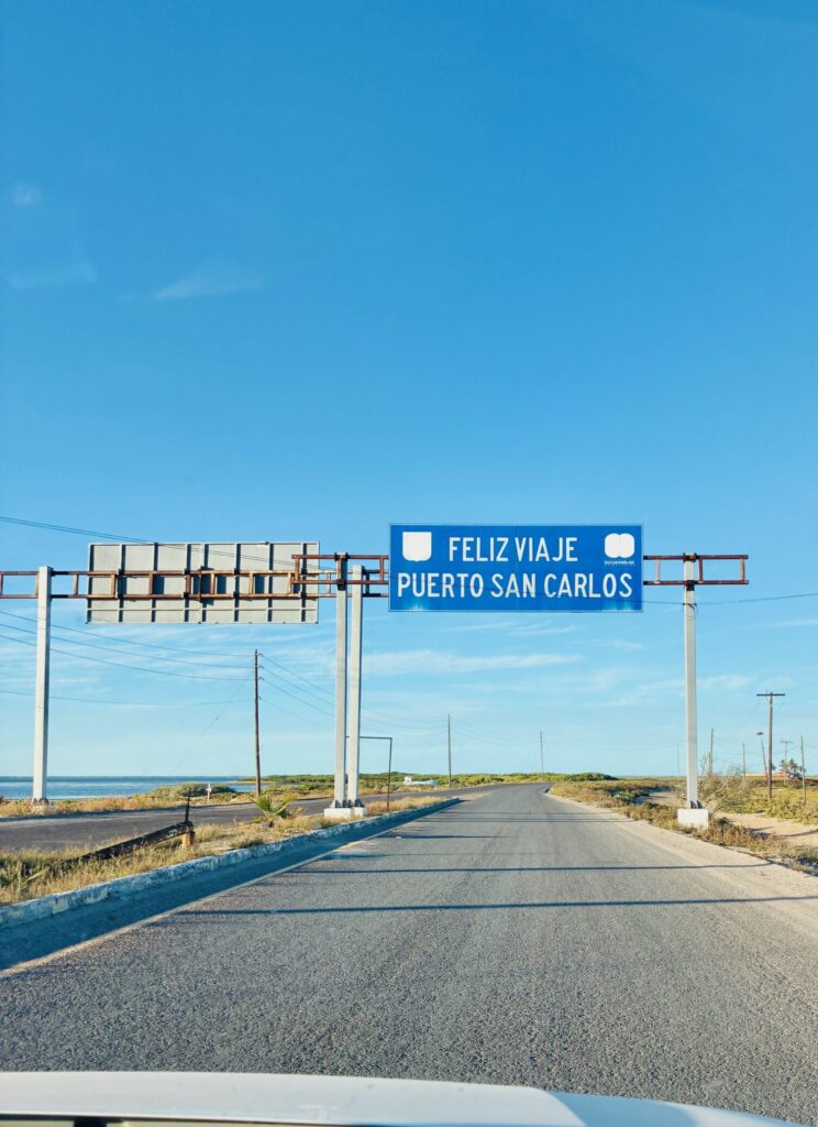 Straßenszene mit blauem Verkehrsschild über der Straße, darauf steht 'FELIZ VIAJE PUERTO SAN CARLOS'