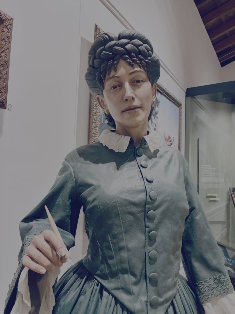 Weibliche Figur mit geflochtenem Haar und historischer Kleidung mit Knöpfen und Spitzenmanschetten, hält einen Stift in der rechten Hand