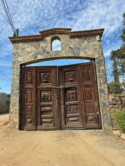 Freistehendes großes Tor aus dunklem Holz mit dekorativen Reliefs, eingebettet in eine Mauer aus unregelmäßigen Natursteinen unter blauem Himmel auf sandigem Weg