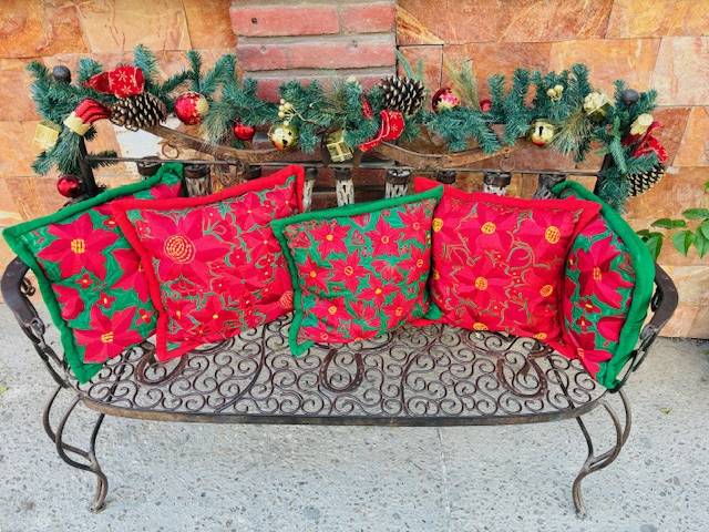 Eiserne Gartenbank mit fünf weihnachtlich dekorierten Kissen in Rot und Grün, dahinter Girlande mit Zapfen und Kugeln.