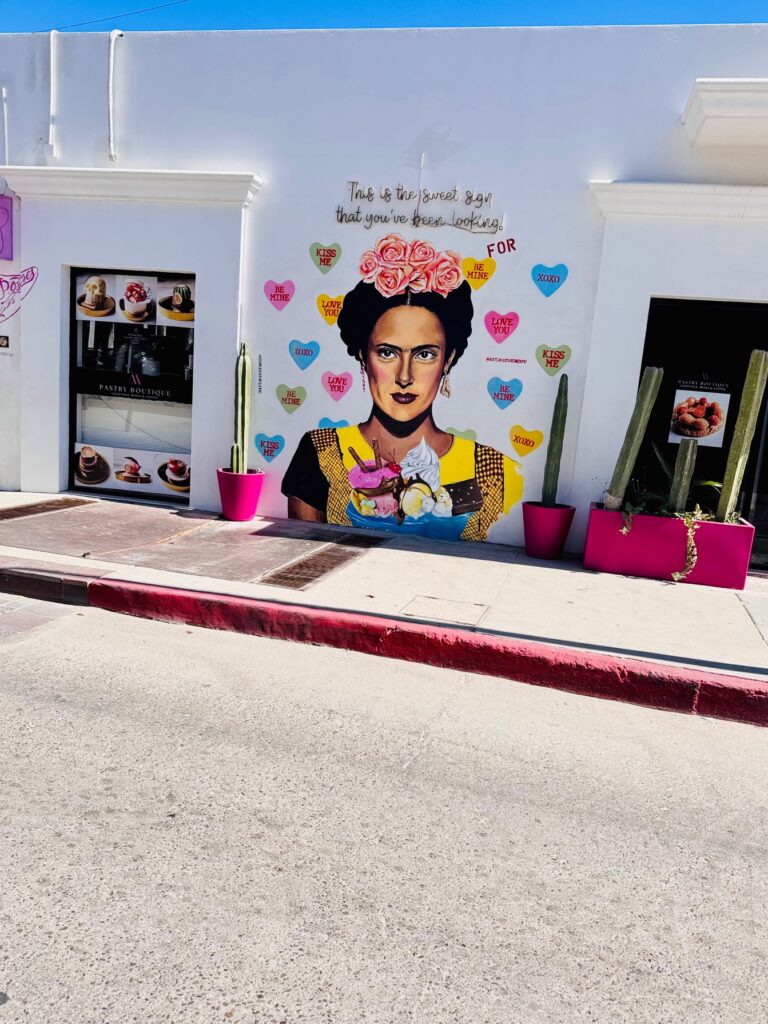 Weiße Wand mit Gemälde einer Frau mit Blumenkranz auf dem Kopf, umgeben von bunten Herzmotiven mit kurzen Texten, zwei große Kakteen in pinken Töpfen daneben unter blauem Himmel