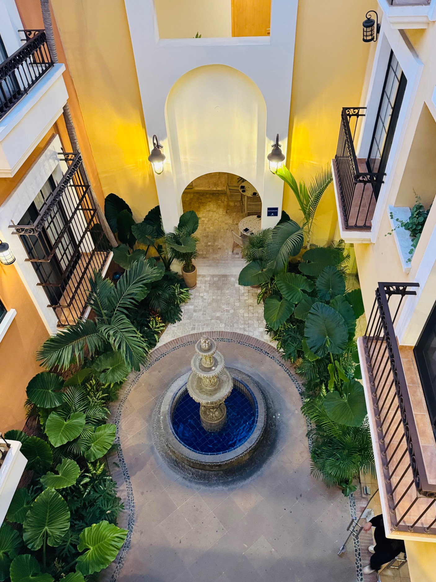 Innenhof mit rundem Brunnen in der Mitte, umgeben von großen grünen Pflanzen und Balkonen an den gelbgestrichenen Wänden